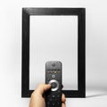 Close-up of male hand holding TV remote against black empty frame on the background of white. Artwork collage concept.