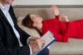 Close-up of a male psychotherapist`s hand writing on a clipboard. A woman in a red dress lies on a couch and talks about