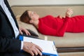 Close-up of a male psychotherapist`s hand writing on a clipboard. A woman in a red dress lies on a couch and talks about