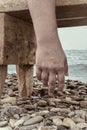 Close up man hand hanging from a beach bench, pebbles and sea on background. Atmosphere of total exhausting, prostrate, solitude,