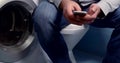 Close-up a man in jeans sits on a closed toilet lid, holds a mobile phone in his hands, leads his fingers on the screen
