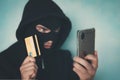 Close up of a man in robbery mask and hood holding the credit card and looking at the smartphone screen. Male criminal arranges a