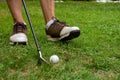 Close-up of man`s feet with golf shoes and out of focus golf club and ball, on grass, horizontal,