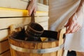 Close-up of a man in a towel scooping water with a ladle from a wooden bucket in a sauna.