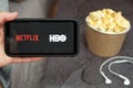 Close up mans hand holding a mobile phone with Netflix and HBO logo with Apple earphones and popcorn box next to him