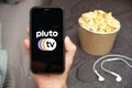 Close up mans hand holding a mobile phone with Pluto tv logo with Apple earphones and popcorn box next to him, Free TV