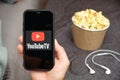 Close up mans hand holding a mobile phone with YouTube TV logo with Apple earphones and popcorn box next to him, Free TV Royalty Free Stock Photo