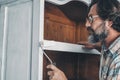 Close up of mature man painting and restoring an old antique wooden cabinet at home with roller and white paint. Recycling and