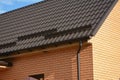 A close-up on a metal brown roof of a brick house with snow stoppers, snow guards, a rain gutter, a downspout, soffit and fascia