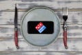 Close up of mobile phone screen with logo lettering of food delivery service dominos pizza on wood table with dish and cutlery Royalty Free Stock Photo