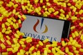 Close up of mobile phone screen with logo lettering of Novartis pharmaceutical company on pile yellow red drug capsules