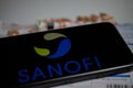 Close up of mobile phone screen with logo lettering of Sanofi pharmaceutical company, blurred pills and lab sheet background