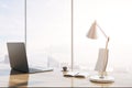 Close up of modern backlit wooden desk with laptop, table lamp, coffee cup and panoramic window with city view in the background.