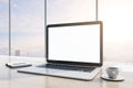 Close up of wooden desktop with blank white mock up laptop screen, supplies, coffee cup and panoramic window with city view Royalty Free Stock Photo