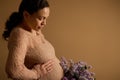 Close-up pregnant woman stroking her tummy, holding bouquet of purple blooming lilacs, isolated beige studio background Royalty Free Stock Photo