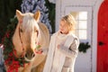 Close-up nice blonde curly kid and adorable pony with festive wreath near the small wooden house and snow-covered trees. New Year Royalty Free Stock Photo