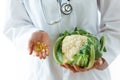 Young nutritionist holding omega pills in one hand and cauliflow in the other hand over white background.