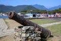 Close-up of an old rusty cannon in front of an open space with grass, colorful boats and rainforest mountains, historic town
