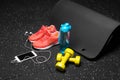 A close-up of orange sport shoes, yellow dumb-bells, pilates mat, blue bottle, and phone with headphones on a black