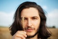 Close-up outdoor portrait of handsome guy with long hair, holding wheat spike.