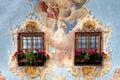 Close up of a painted facade with two wooden windows and geraniums on the windowsill