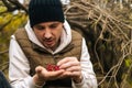 Close-up of pensive hunger tourist man sitting under tree in forest and holding wild red berries in hands, looking at