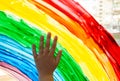 Close-up photo of child`s hand touch painting rainbow on window. Family life background. Image of kids leisure at home