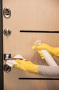 Close up entrance door knob being cleaned with sanitizer spray by female hands in yellow latex gloves, selective focus