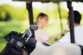 Close up photo. Golf clubs in the foreground, man with a woman in a golf cart on a background