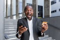 Happy african american man standing outside. He holds a phone and a credit card, rejoices, shouts Royalty Free Stock Photo