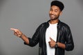 Close-up photo of happy young afro american man in leather jacket pointing with figer while showing thumb up gesture, looking Royalty Free Stock Photo