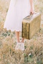 The close-up photo of the legs of the bride dressed in the knee-length dress and carrying the vintage suitcase in the