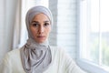 Close-up photo. Portrait of a young Muslim woman in a hijab standing by the window at home, confidently and seriously Royalty Free Stock Photo