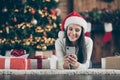 Close up photo of positive girl in santa claus hat lie on carpet use smartphone send christmas congratulation search