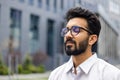 Close-up photo of a relaxed young Indian man wearing glasses and a white shirt sitting outside a business center with Royalty Free Stock Photo