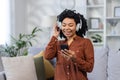 Close-up photo of a smiling young African-American woman sitting on the sofa at home, wearing headphones and using a Royalty Free Stock Photo