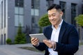 Close-up photo of a smiling young Asian man sitting outside in a suit and headphones, holding a tablet and talking on a Royalty Free Stock Photo