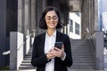 Close-up photo of a smiling young business woman in a suit and glasses standing outside an office building and using a Royalty Free Stock Photo