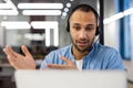 Close-up photo of a smiling young hispanic man in a headset sitting in the office in front of a laptop and talking on a Royalty Free Stock Photo