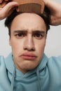 Close-up photo. A surprised young man combs his hair in a light light blue hoodie,with a comb in his hand combing a