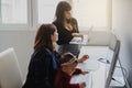 photo of two women in office outfit working on computers and discussing a report; one of them is with a baby girl in her arms