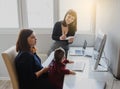 photo of two women in office outfit working on computers and discussing a report; one of them is with a baby girl in her arms