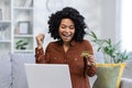 Close-up photo of young African American woman sitting on sofa at home with laptop, looking at credit card, happy and Royalty Free Stock Photo