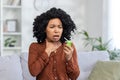 Close-up photo of a young African-American woman treating her throat with a medical spray at home, sitting on the couch Royalty Free Stock Photo