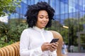 Close-up photo of a young African-American woman in a white shirt sitting on a bench outside and smiling while using a Royalty Free Stock Photo