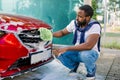 Close up photo of young African man cleaning radiator grille of luxury red car with foam and green brush mitten. Carwash