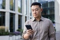 Close-up photo of a young Asian male businessman standing outside an office center with a phone in his hands, waiting Royalty Free Stock Photo