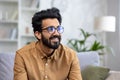 Close-up photo of a young Indian man in glasses sitting on the couch at home and looking to the side with a smile Royalty Free Stock Photo