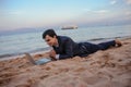 Photo of a young man in suit with laptop lies on the beach and talking to someone on computer Royalty Free Stock Photo