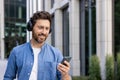 Close-up photo of a young smiling man standing on a city street wearing headphones and using a mobile phone. Listens to Royalty Free Stock Photo